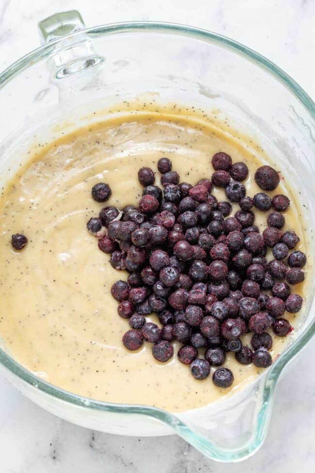 A clear glass mixing bowl filled with muffin batter and blueberries.