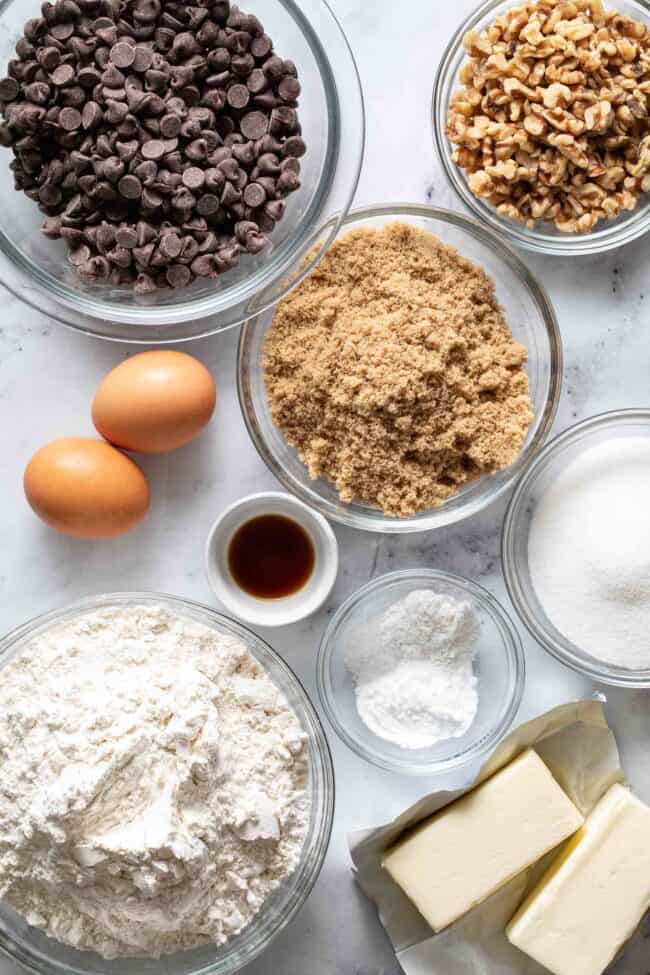 Glass mixing bowls filled with flour, brown sugar, white sugar, chocolate chips and walnuts sit next to 2 sticks of butter and 2 eggs.