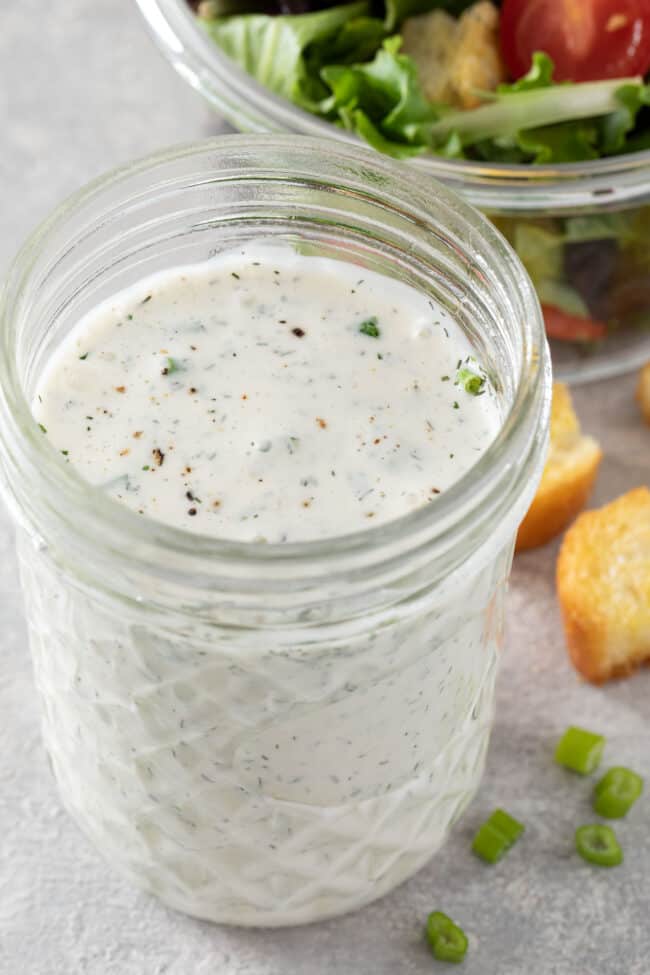 A clear glass mason jar filled with Vegan Ranch Dressing. A clear glass bowl filled with mixed salad sits next to the mason jar.