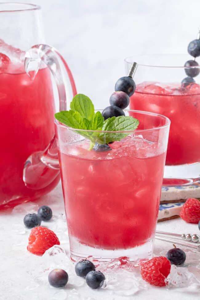 Two clear glasses and a clear glass pitcher filled with iced blueberry green tea. Fresh berries are scattered around the glasses.