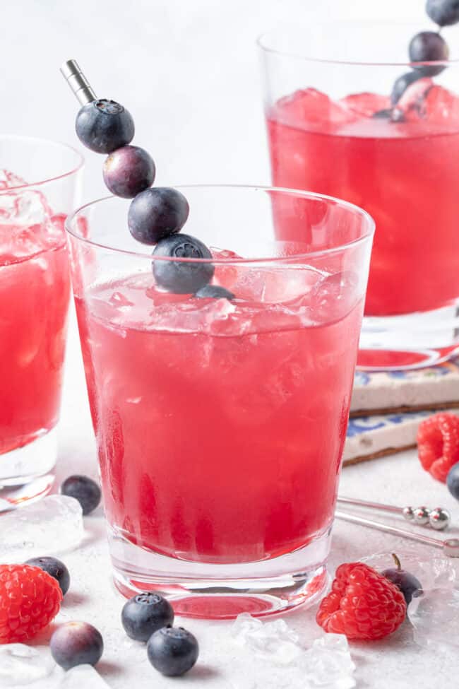 Three clear glasses filled with bluish liquid with berries scattered around the glasses.