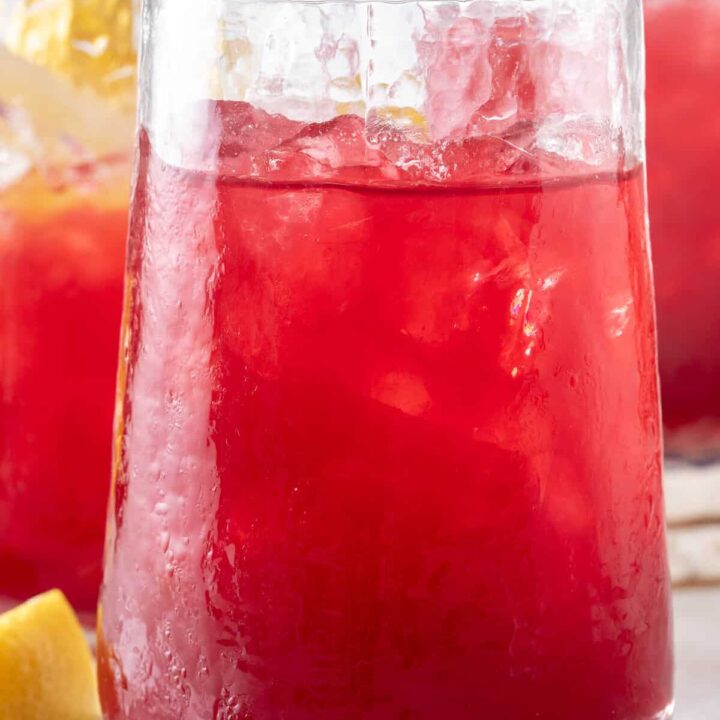 Three clear drinking glasses filled with Blueberry Lemonade. The glasses are sitting on a wooden cutting board with blueberries and lemon wedges.