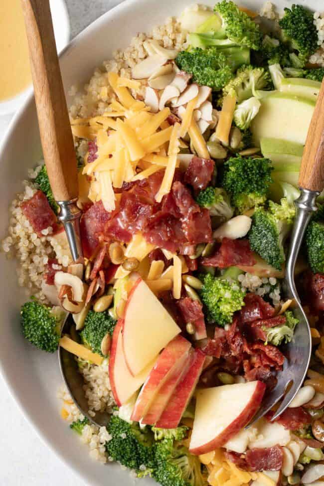 A white bowl filled with Broccoli Cheddar Quinoa Salad. Serving spoons rest inside the salad bowl.