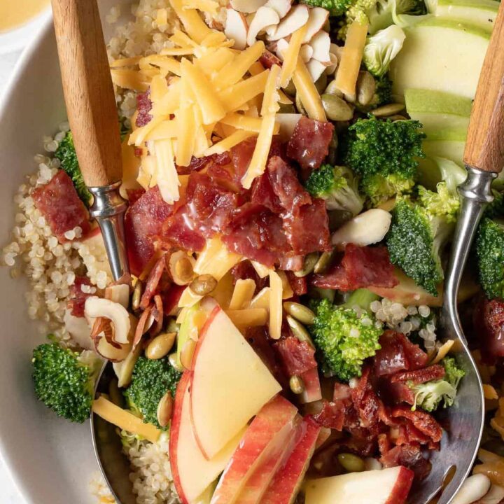 A white bowl filled with Broccoli Cheddar Quinoa salad. Salad spoons rest in the bowl.