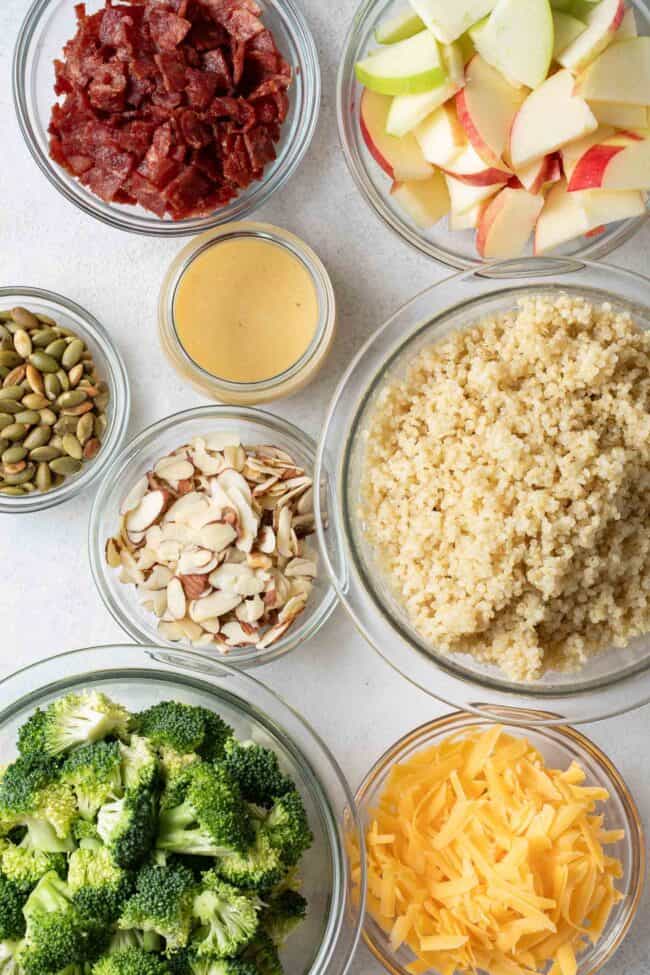 Clear glass mixing bowls filled with quinoa, broccoli, grated cheese, nuts, seeds, chopped apple and dressing.