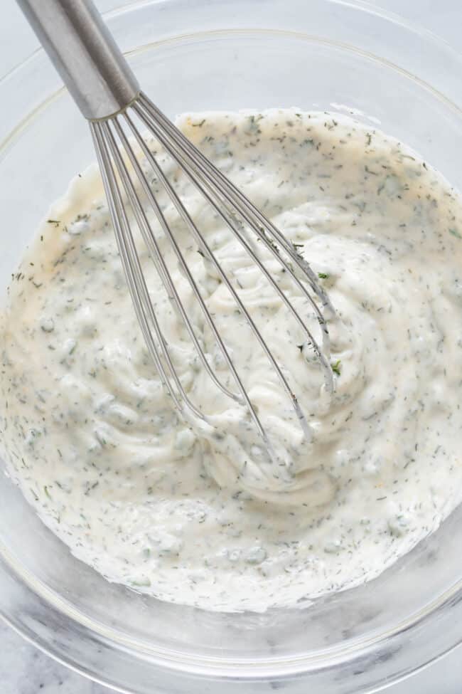 A clear glass mixing bowl with mayonnaise and herbs whisked together with a wire whisk.