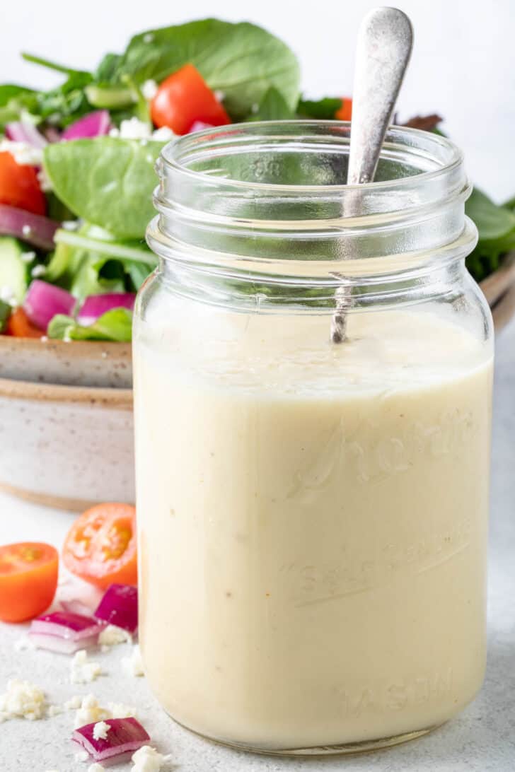 A clear mason jar filled with champagne vinaigrette. A tan colored bowl filled with salad sits behind the jar.