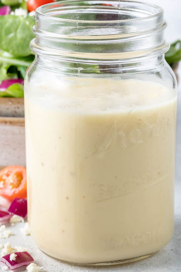 A clear glass mason jar filled with champagne vinaigrette. A tan colored bowl filled with salad sits behind the jar.