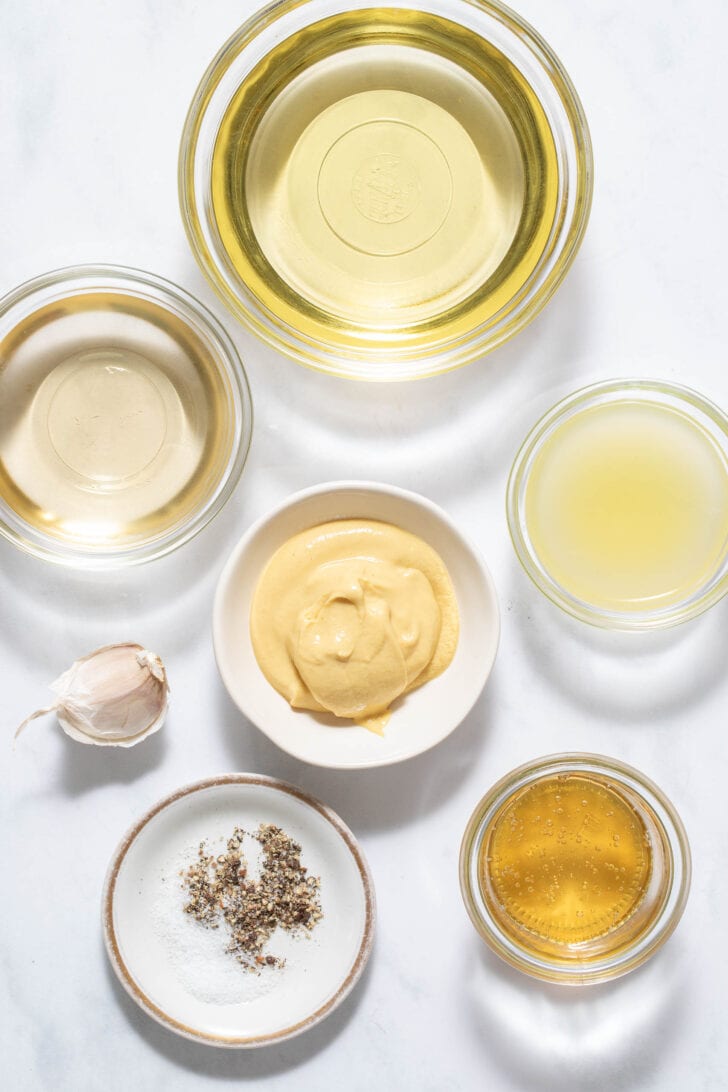 Six clear glass bowls filled with olive oil. vinegar, Dijon mustard, honey, lemon juice and salt and pepper.