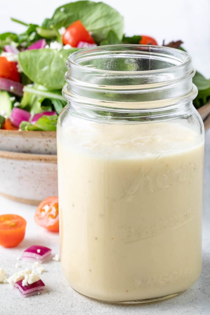 A clear glass mason jar filled with champagne vinaigrette. A tan colored bowl filled with salad sits behind the jar.
