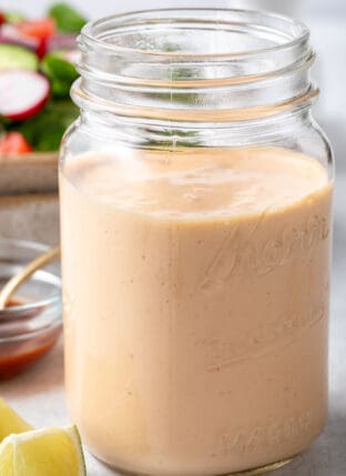 A clear glass mason jar filled with Chipotle Ranch Dressing, A bowl filled with salad sits behind the jar.