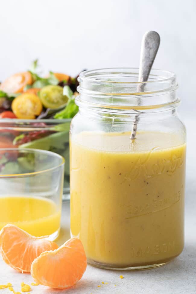 A mason jar filled with citrus vinaigrette with a clear glass bowl filled with salad behind it.