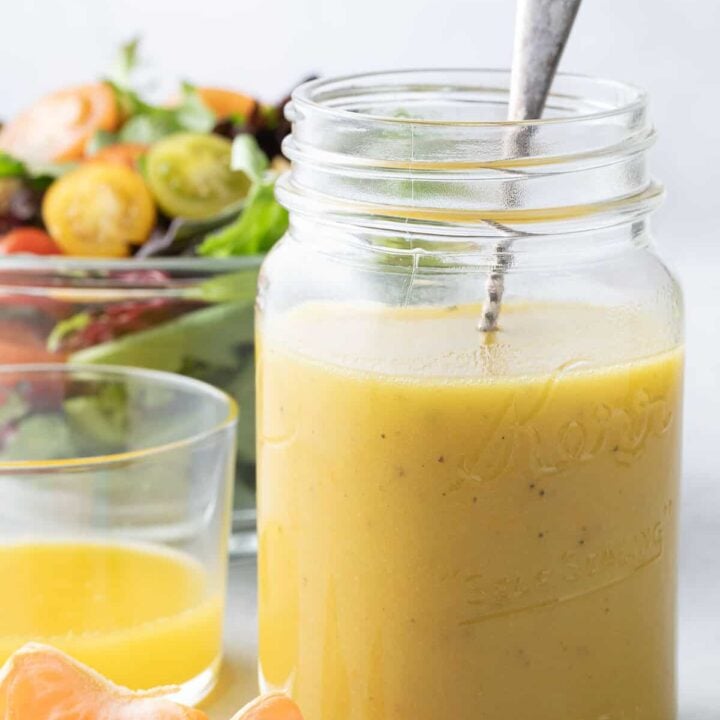 A clear mason jar filled with Citrus Vinaigrette. A clear glass bowl filled with salad sits behind the jar.