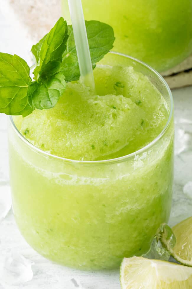 A clear glass filled with a green drink. A mint sprig and straw are poked into the drink.