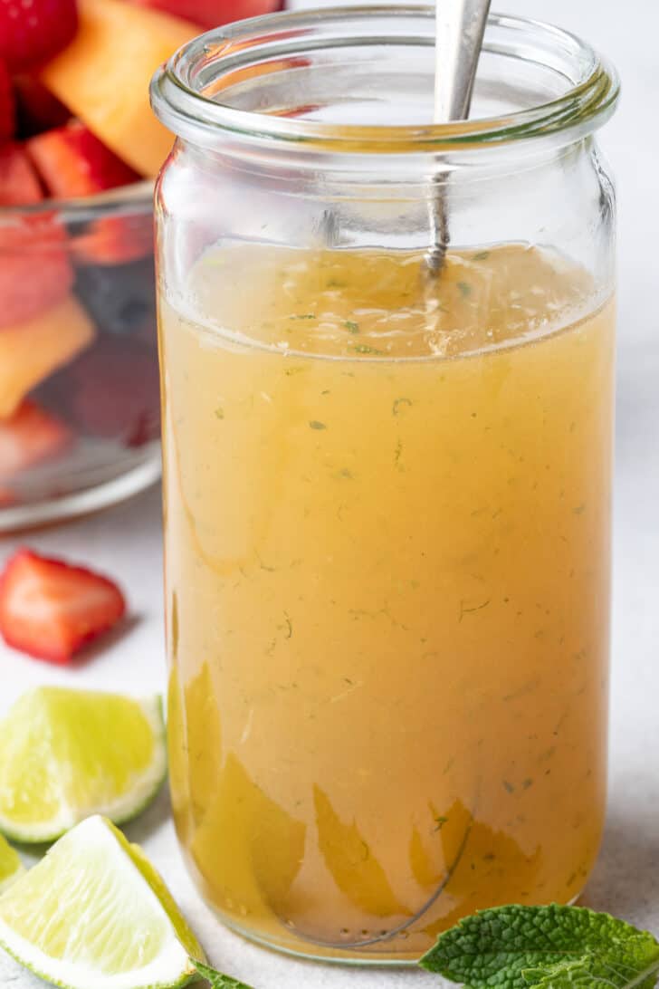 A clear glass mason jar filled with lime juice and honey. A clear glass bowl filled with fruit sits next to the jar.
