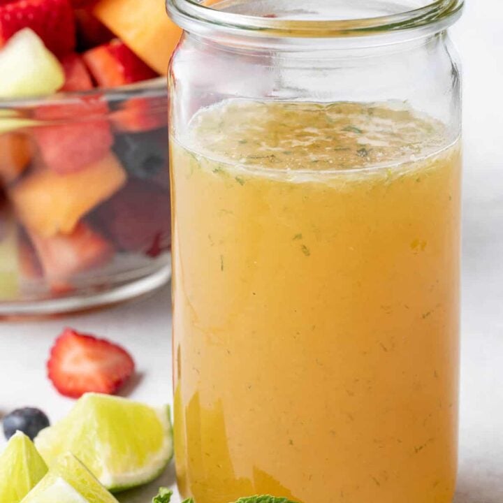 A clear glass mason jar filled with Fruit Salad Dressing. A clear glass bowl filled with fruit salad sits next to the jar.