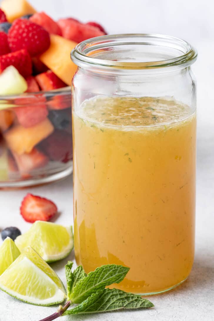 A clear glass mason jar filled with lime Fruit Salad Dressing. A clear glass bowl filled with fruit salad sits next to the jar.