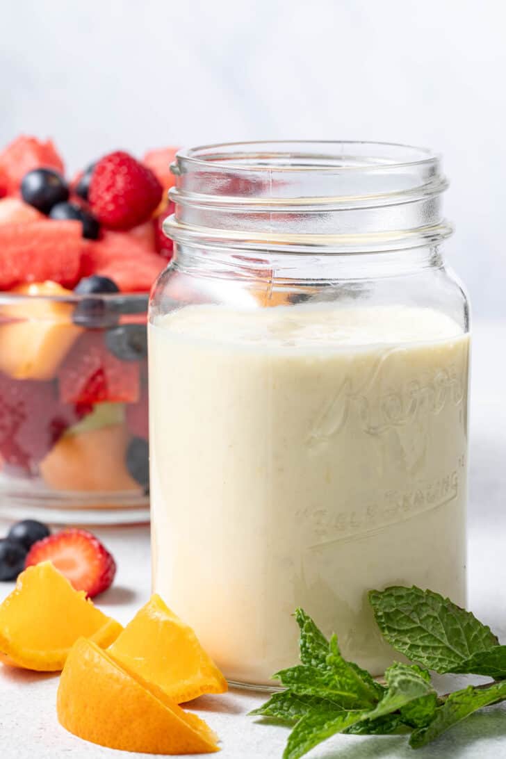 A clear glass mason jar filled with creamy Greek Yogurt Fruit Salad Dressing. A clear glass bowl filled with fruit salad sits next to the jar.