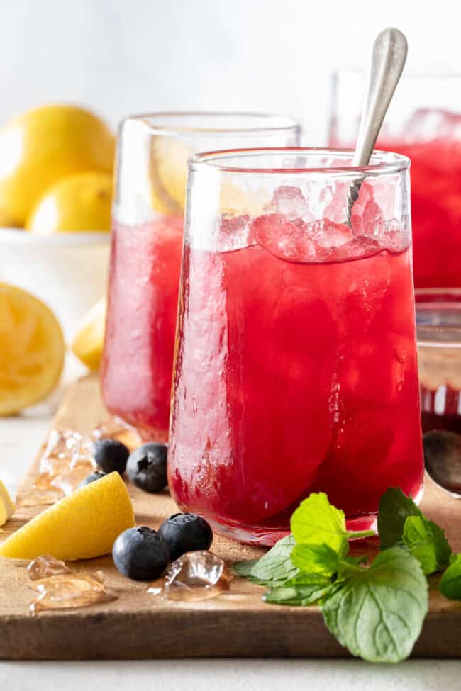 Three clear drinking glasses filled with Blueberry Lemonade. The glasses are sitting on a wooden cutting board with blueberries and lemon wedges.