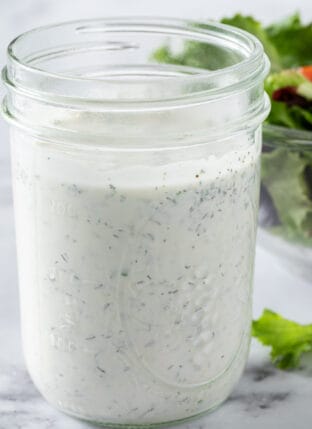 A mason jar filled with ranch dressing. A clear glass bowl filled with salad sits next to the jar.