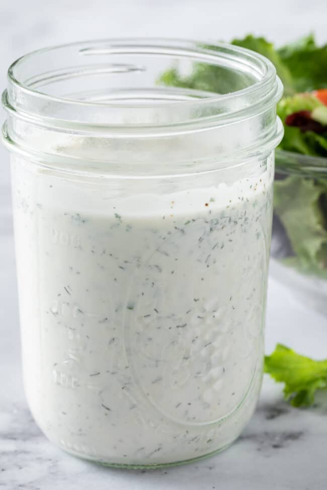 A clear glass mason jar filled with homemade ranch dressing. A clear glass bowl filled with salad sits next to the jar.