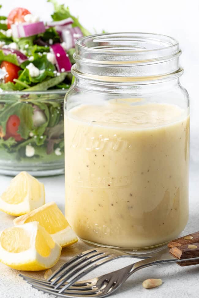 A clear glass mason jar filled with Honey Lemon Vinaigrette. A clear glass bowl filled with salad sits next to the jar.