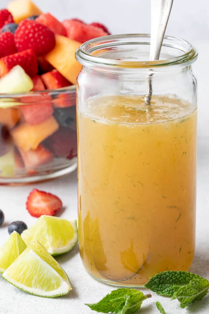 A clear mason jar filled with honey lime dressing for fruit salad. A clear glass bowl filled with fruit salad sits next to the jar.