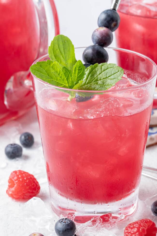 Two glasses and a clear glass pitcher filled with iced blueberry iced tea.