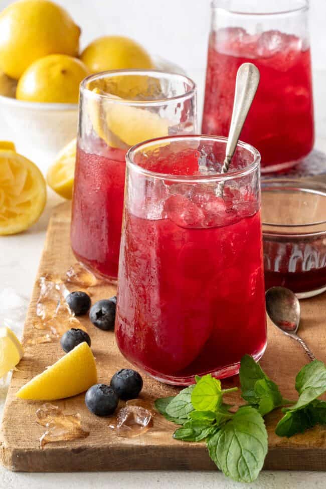 Three clear drinking glasses filled with Blueberry Lemonade. The glasses are sitting on a wooden cutting board with blueberries and lemon wedges.