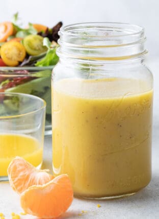 A clear glass mason jar filled with citrus vinaigrette. A clear glass bowl filled with salad is behind the mason jar.