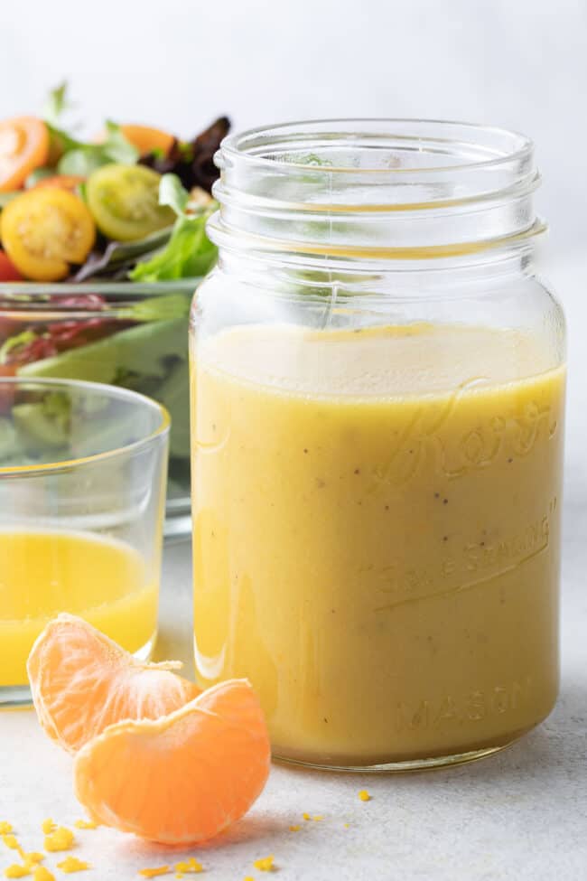A clear glass mason jar filled with citrus vinaigrette. A clear glass bowl filled with salad sits behind the jar.