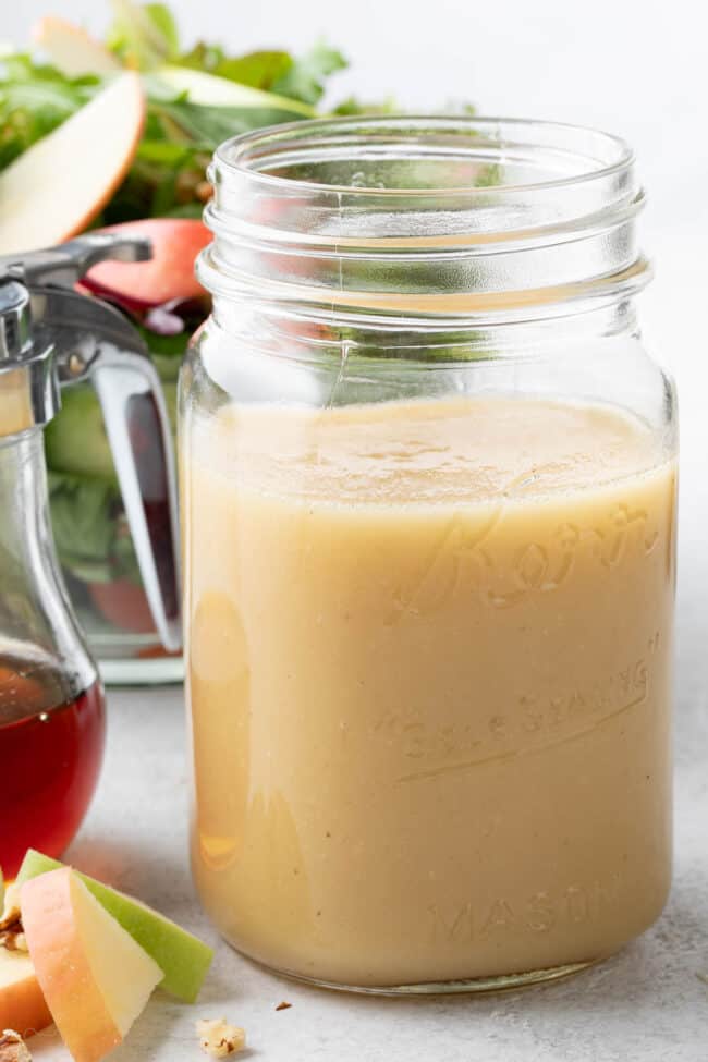 Clear glass mason jar filled with maple vinaigrette. A salad in a clear bowl sits behind the mason jar.