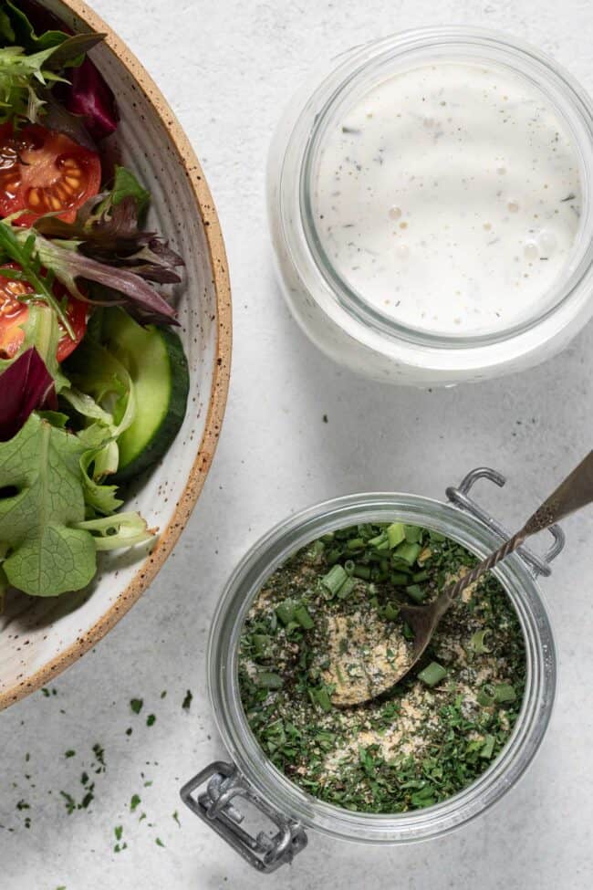 A small mason jar filled with Ranch Dressing Seasoning. Another mason jar is filled with ranch dressing and a bowl filled with salad sits next to the jars.