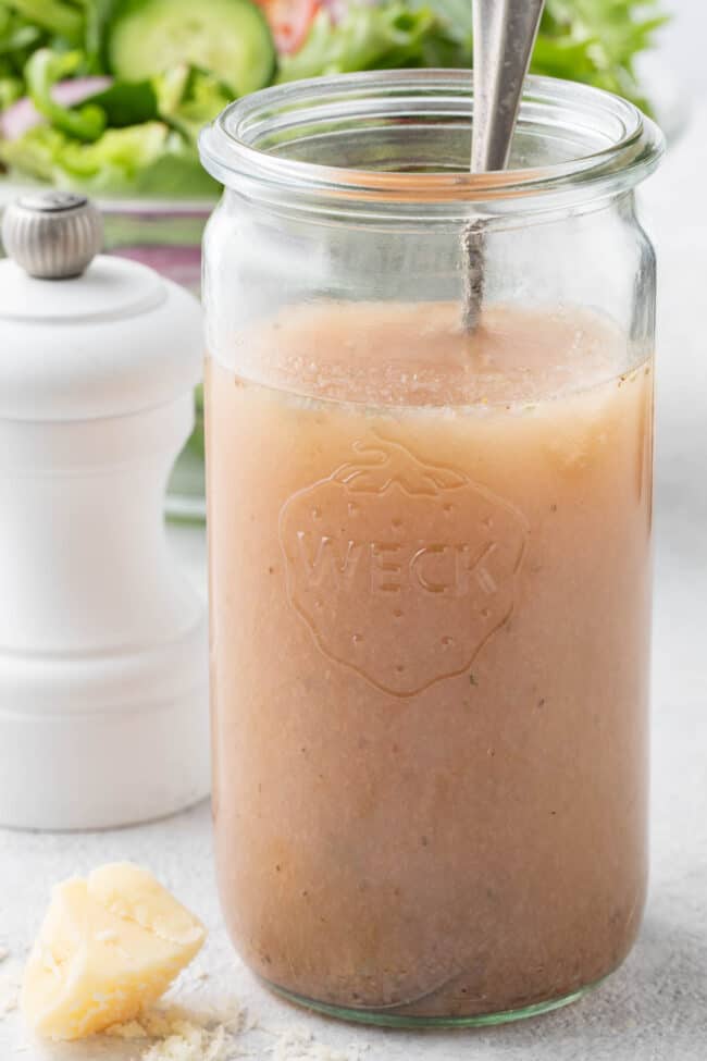 A clear glass mason jar filled with red wine vinaigrette dressing. A clear glass bowl filled with salad sits behind the jar.