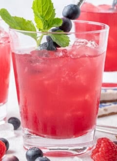 Three clear glasses filled with Iced Blueberry Green Tea. Blueberries and raspberries are scattered around the glasses.