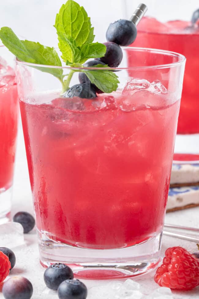 Three clear glasses filled with iced blueberry green tea. Blueberries and raspberries are scattered around the glasses.