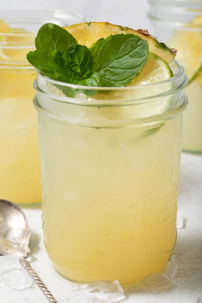 A clear glass mason jar filled with Iced Pineapple Mint Green Tea. The glass is garnished with pineapple, mint and lime slices.