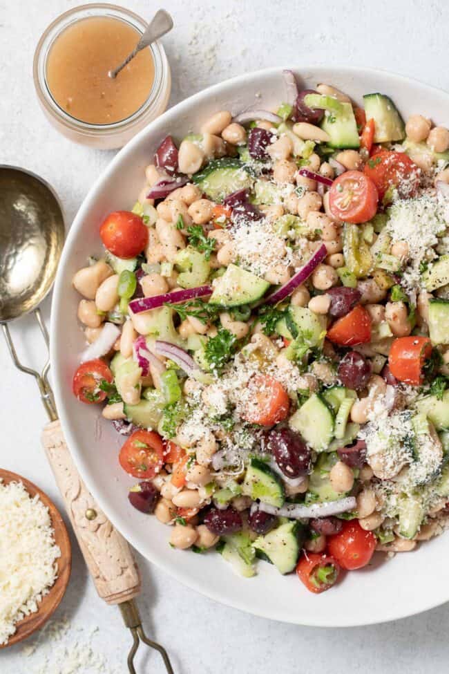 A large white salad bowl filled with white bean salad mixed with vegetables. A large salad spoon sits next to the bowl.