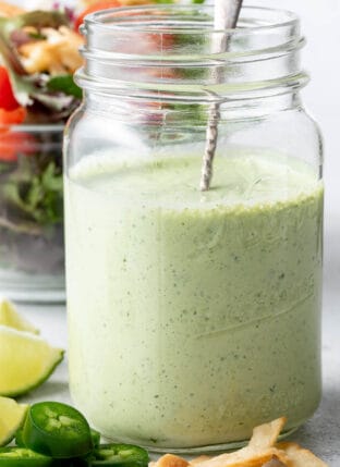 A clear glass mason jar filled with Jalapeno Ranch Dressing. A clear glass bowl filled with salad sits next to the jar.