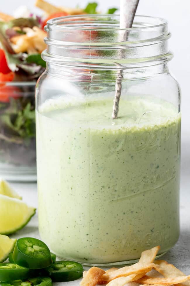 A clear glass mason jar filled with Jalapeno Ranch Dressing. A clear glass bowl filled with salad sits next to the jar.