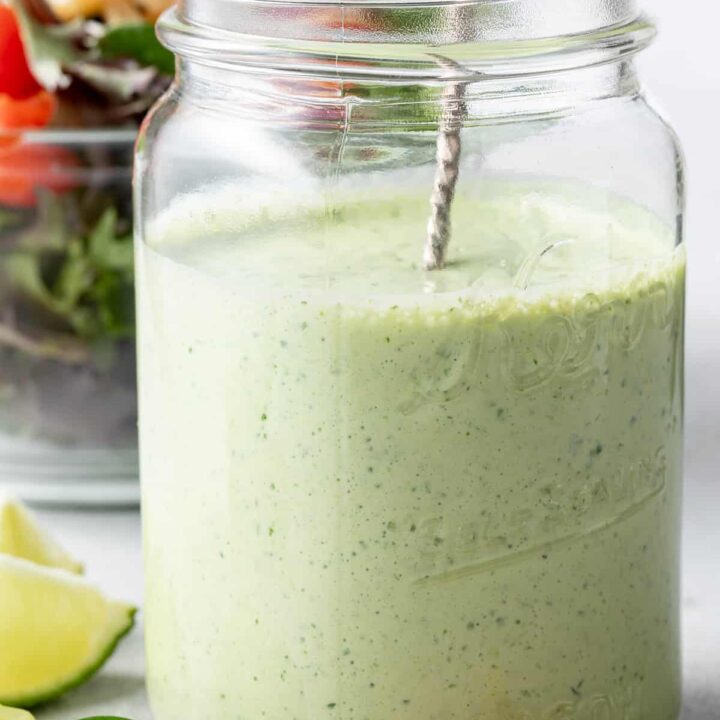 A clear glass mason jar filled with Jalapeno Ranch Dressing. A clear glass bowl filled with salad sits next to the jar.