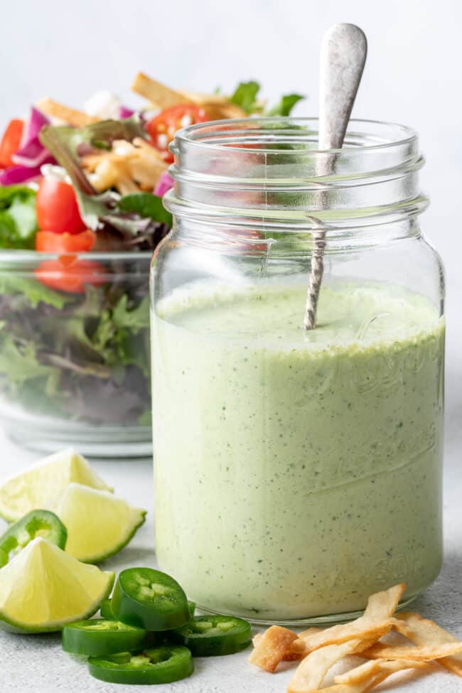 Clear mason jar filled with Jalapeno Ranch Dressing. A clear glass bowl filled with salad sits next to the jar.