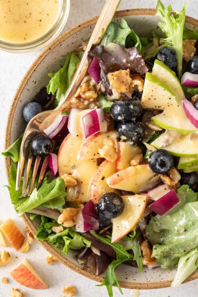 A tan colored bowl filled with a green salad tossed with apples and blueberries.