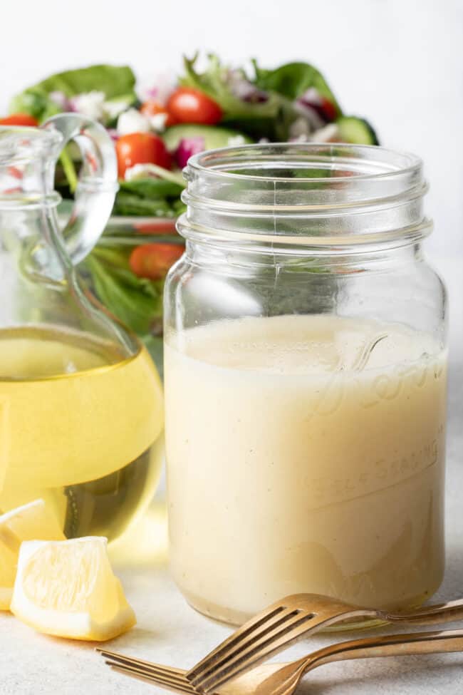 A clear glass mason jar filled with lemon olive oil dressing sits next to a clear glass bowl filled with salad.