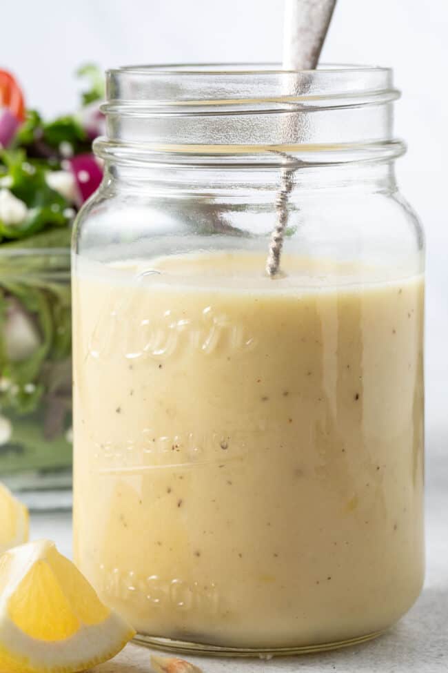 A clear glass mason jar filled with Honey Lemon Vinaigrette. A clear glass bowl filled with salad sits next to the jar.