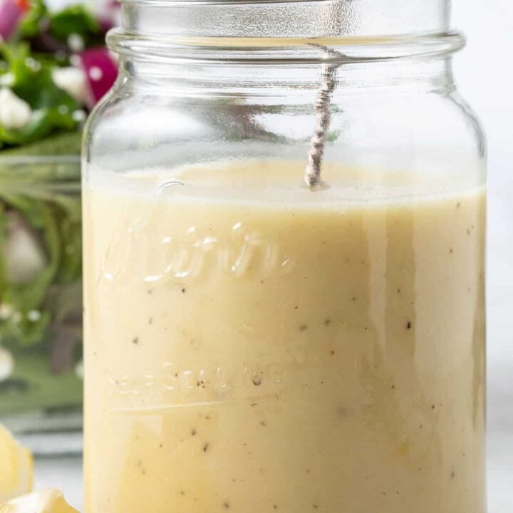 A clear glass mason jar filled with Honey Lemon Vinaigrette. A clear glass bowl filled with salad sits next to the jar.