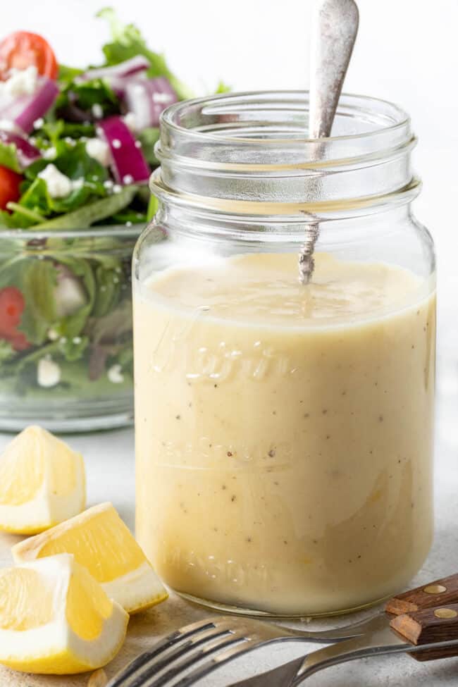 A clear glass mason jar filled with Honey Lemon Vinaigrette. A clear glass bowl filled with salad sits next to the jar.