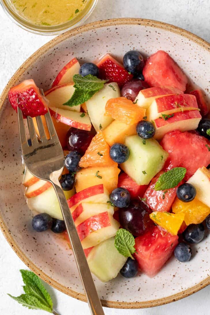A tan colored bowl filled with fruit salad. A gold fork rests in the bowl.