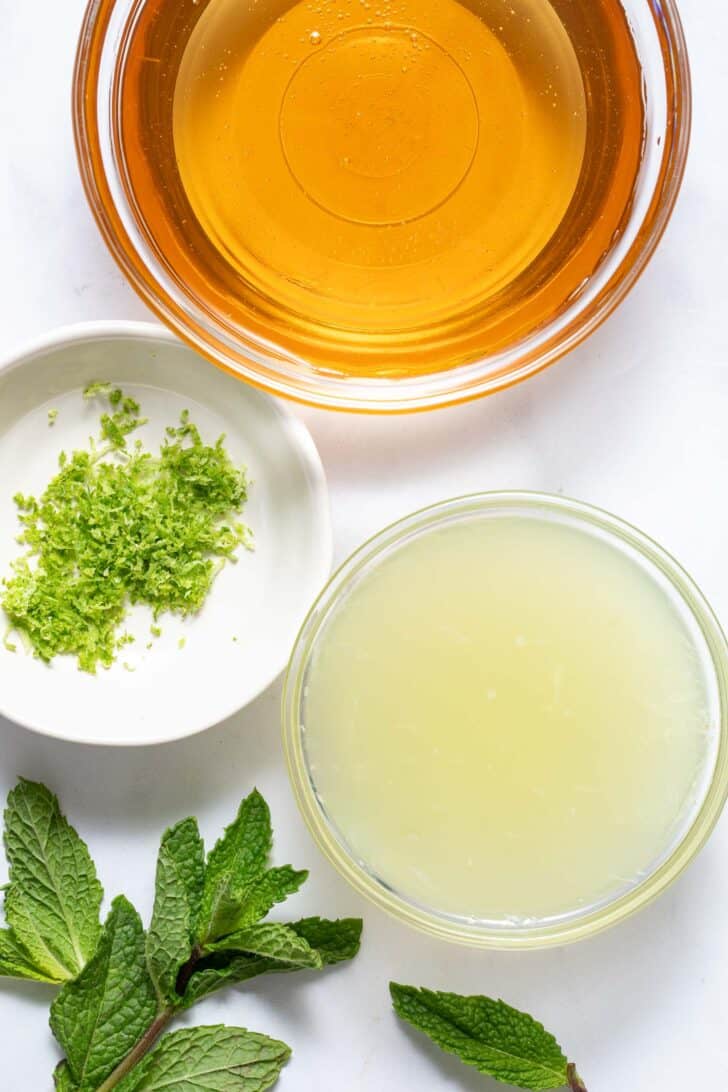 Three small bowls filled with honey, lime juice and lime zest. A sprig of fresh mint sits next to the bowls.