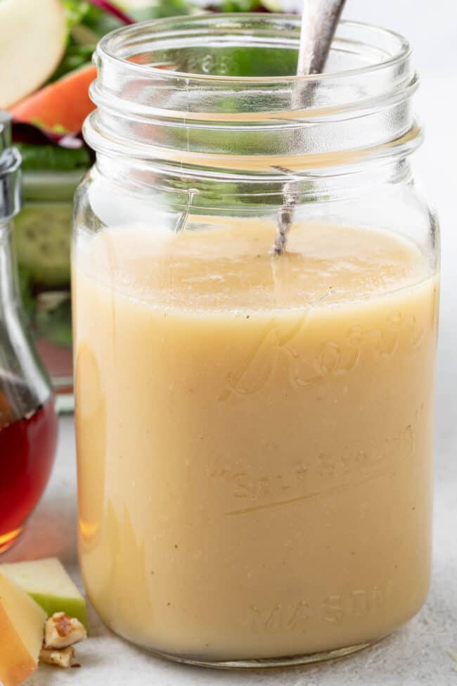 A clear mason jar filled with maple vinaigrette. A salad in a clear glass bowl sits behind the jar.